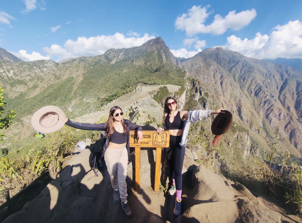 Dos mujeres en la cima de Huayna Picchu, con un letrero, mostrando el desafío de un tour Valle Sagrado y Machu Picchu de 2 días y una noche